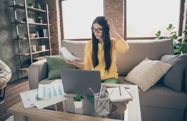 Woman on couch behind laptop with document in her hands